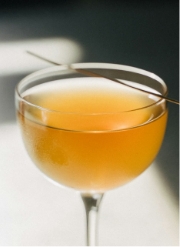 A coupe glass filled with an amber-colored cocktail, garnished with a thin metal stick, sits in natural light on a white surface.